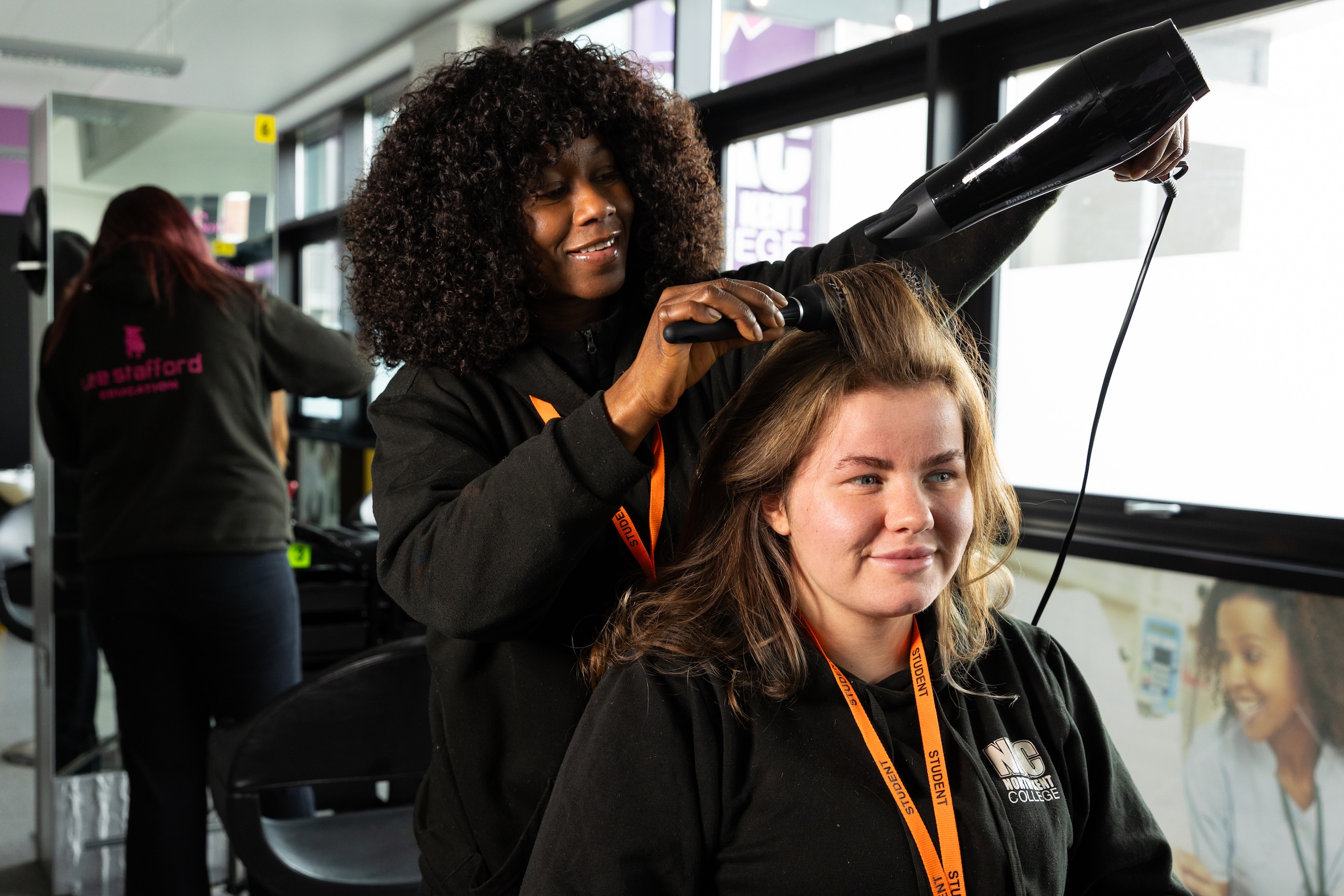 A hairdressing student standing behind a seated student drying her hair